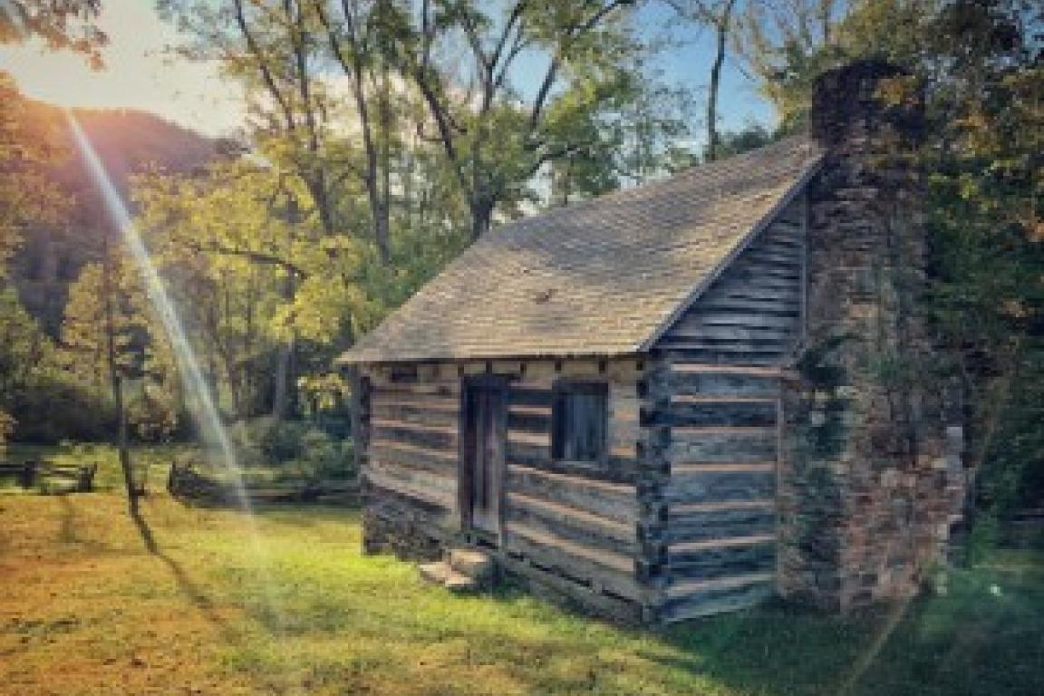 The interpreted slave dwellings on Vance Birthplace. Wooden house with a brick chimney on a field, with trees and sunlight surrounding it
