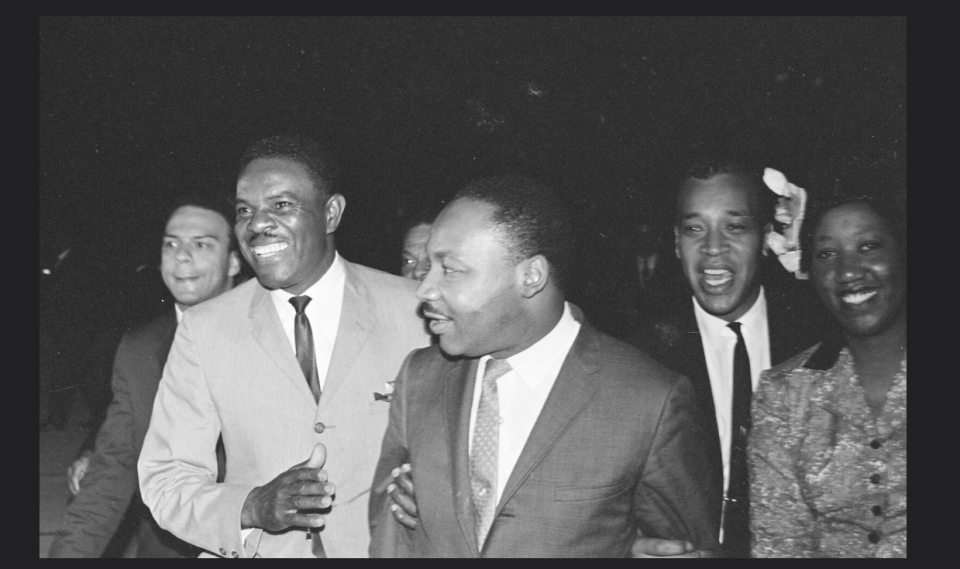 Golden Frinks standing, smiling, arm in arm with Dr. Martin Luther King Jr. Four other people surround them.