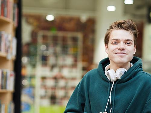  A young person wearing a green hoodie and white headphones in a library or bookstore, with shelves of books behind them.