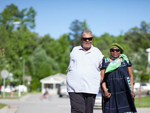  Two people walk hand‑in‑hand along a sunny outdoor path, wearing sunglasses and casual clothing with trees and a white building in the background.