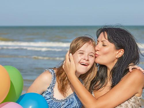 Two people on a beach with ocean waves behind them, smiling as one gently kisses the other’s cheek while colorful balloons float in the foreground.
