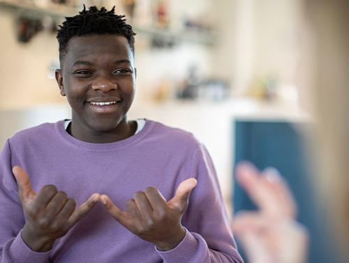  A person in a light purple sweater smiles while communicating with someone whose hands are visible in the foreground, holding a conversation in sign language in a bright indoor setting.