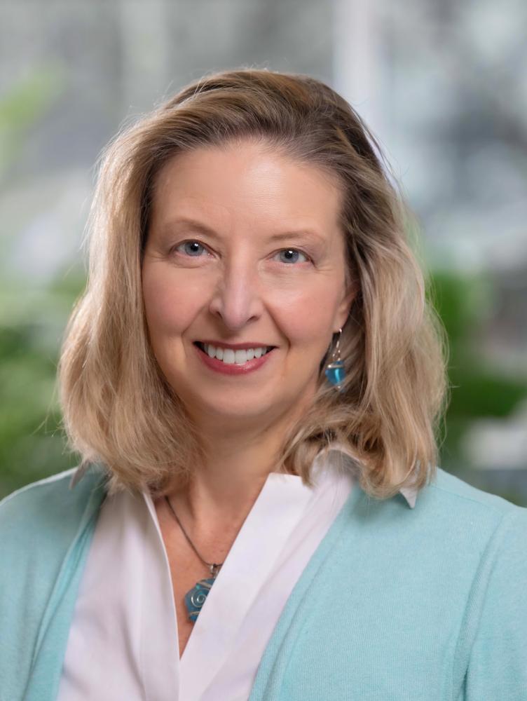 Close up of woman with shoulder-length light brown hair and aqua sweater standing in front of plants
