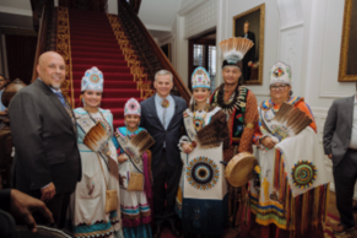 Group of seven people standing in front of a grand staircase; six are in Native American regalia, Gov. Stein is in middle