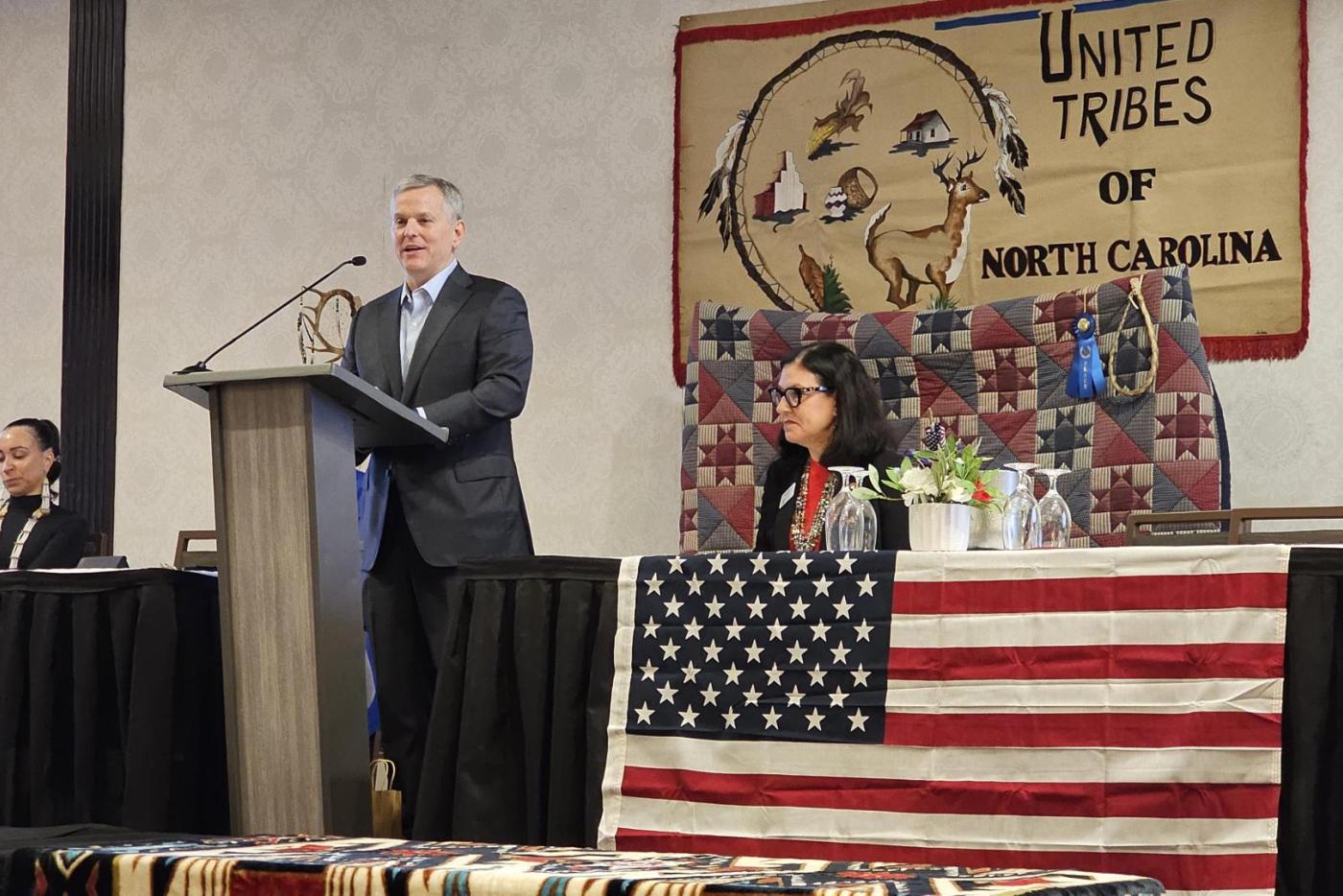 man at podium on stage. speakers seated at tables on either side of him. Tables draped with American flags