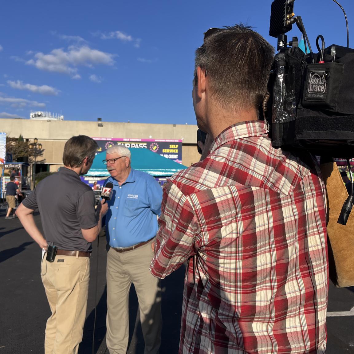 A local t.v. meteorologist interviews Commissioner Steve Troxler as a videographer points a t.v. camera at them during a live report on the fairgrounds