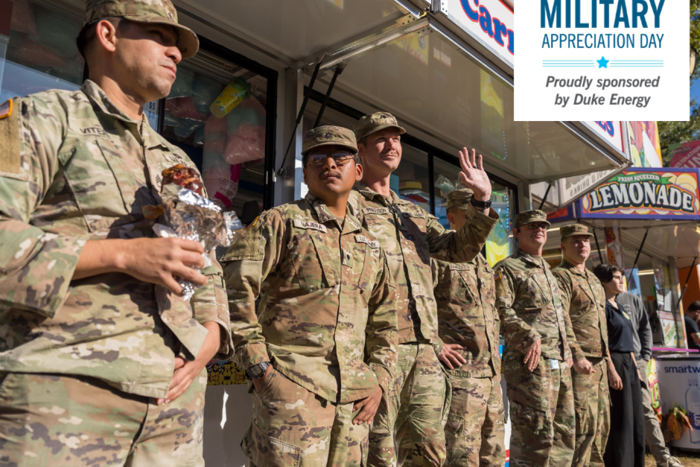 Several uniformed members of the military stand in front of a concession stand, smiling and waving.