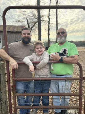Two men and a boy stand behind a fence on a farm.