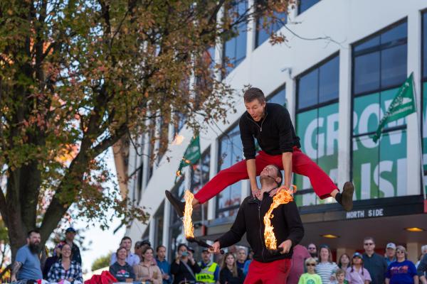 Guy in red trousers jumping over the shoulders of another man holding batons with fire. 