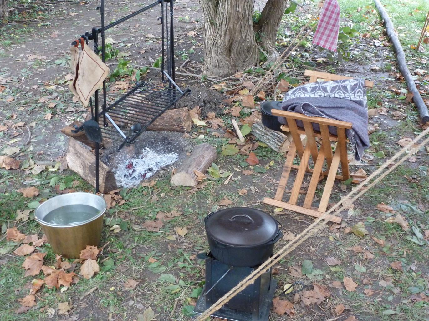 Close up of campsite items, including fire, cooking rack, seat, and water bowl.