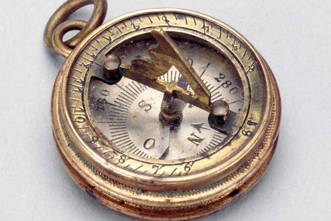 An antique brass compass featuring a sundial raised over the face, photographed over a light background.