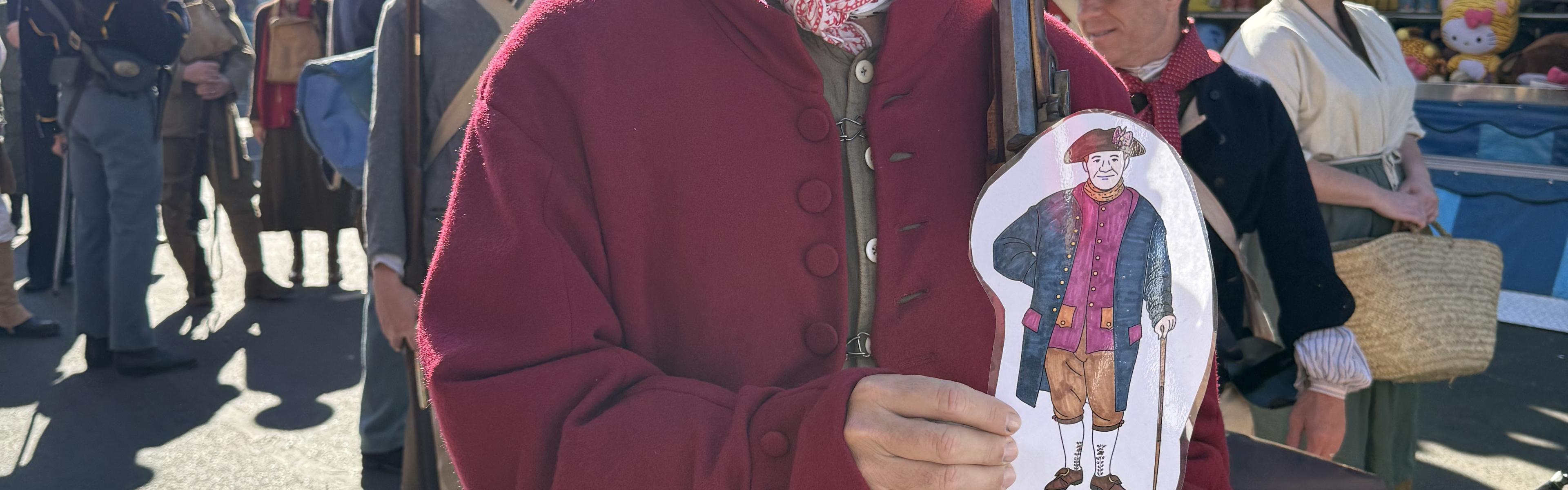 A person dressed in Revolutionary War–era clothing holds a musket and a paper cutout of “Flat Cornelius” while standing at an outdoor fair. Behind them, other reenactors in colonial attire gather near colorful carnival booths and hanging plush prizes.