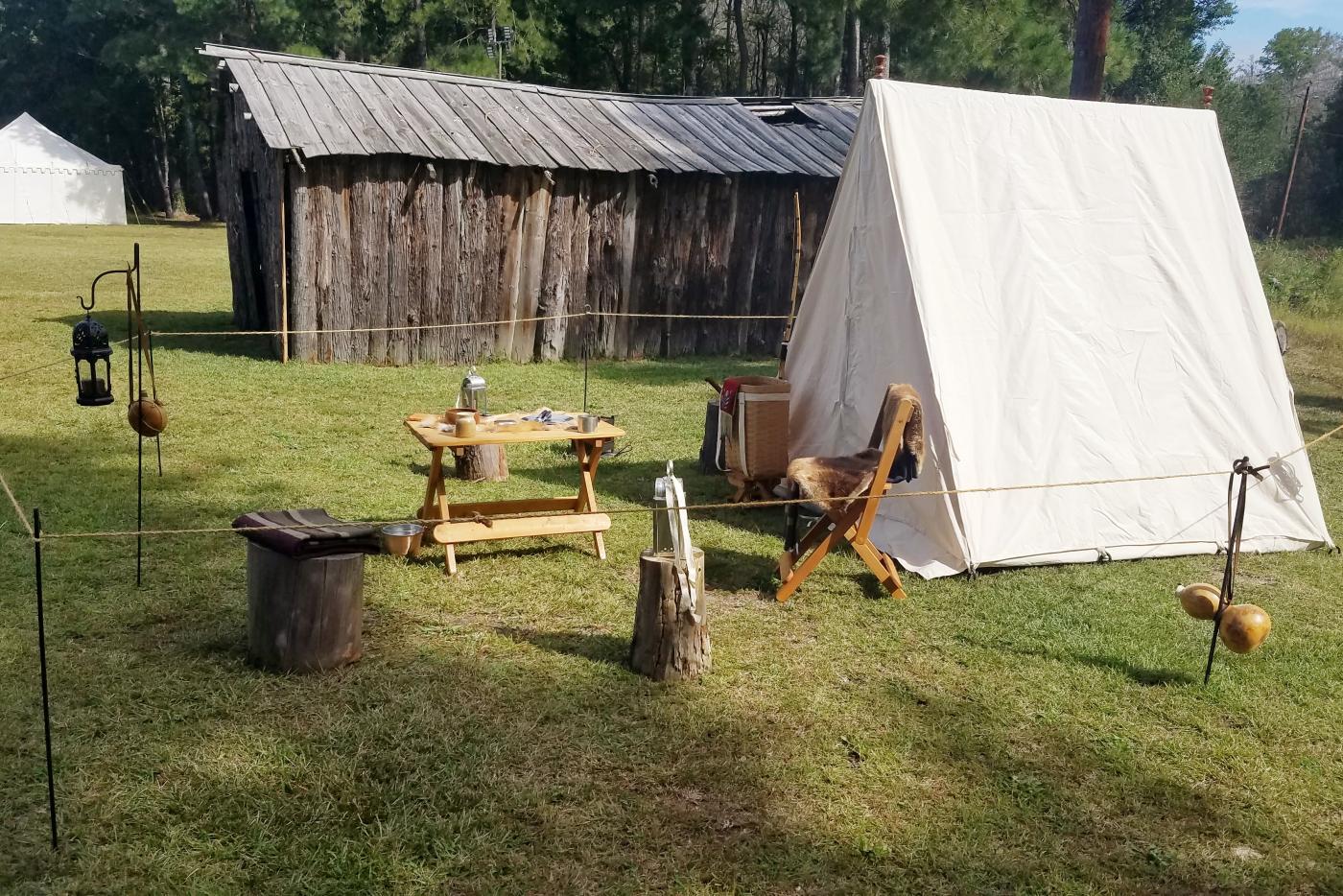 Historic campsite including a white tent, table, and fur covered chair. On grass. Wooden structure in the background.