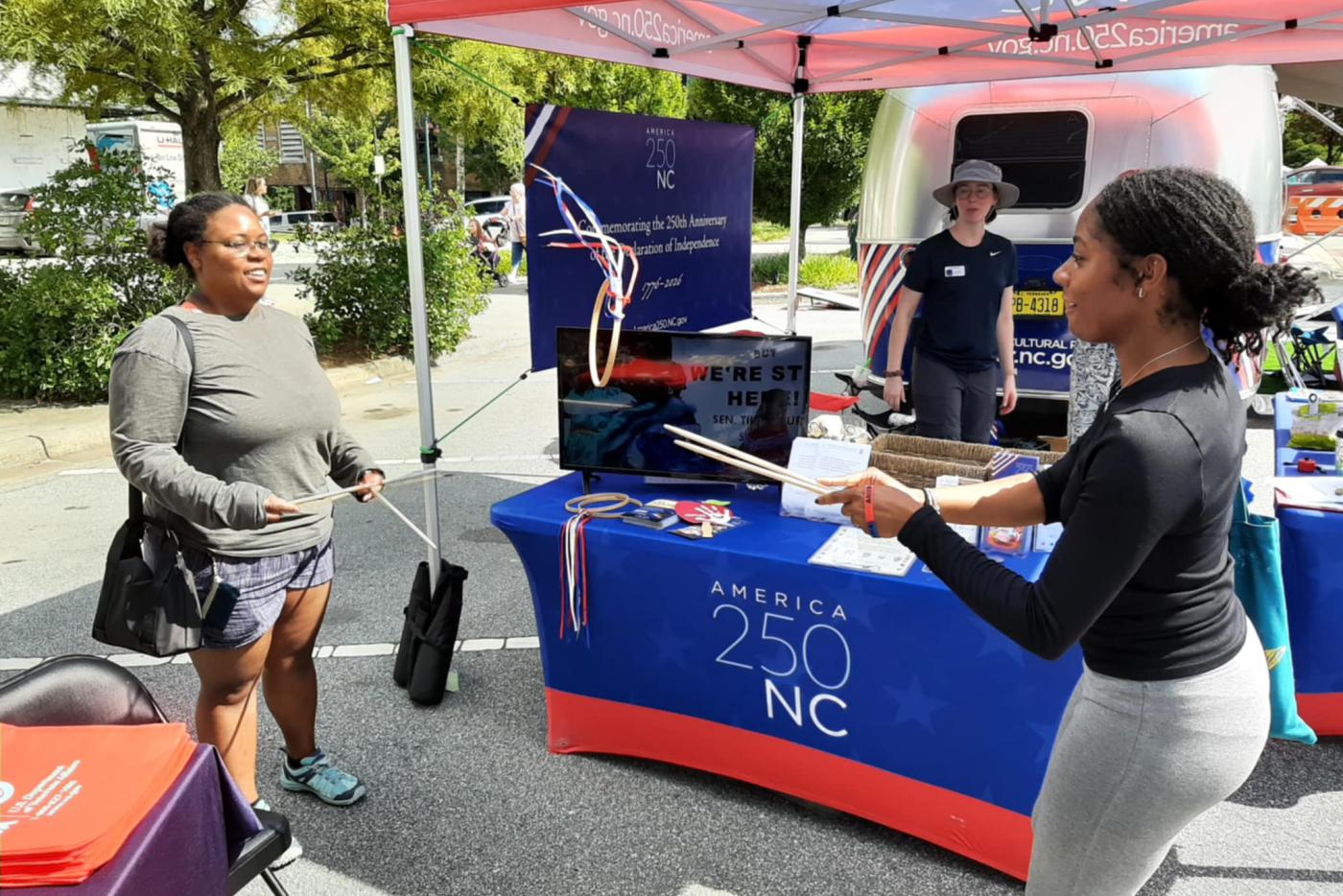 Two women play the historic game of Graces with wooden sticks and hoops at the America 250 NC booth during an outdoor event. A staff member stands nearby under a red tent with informational displays and materials on the table.