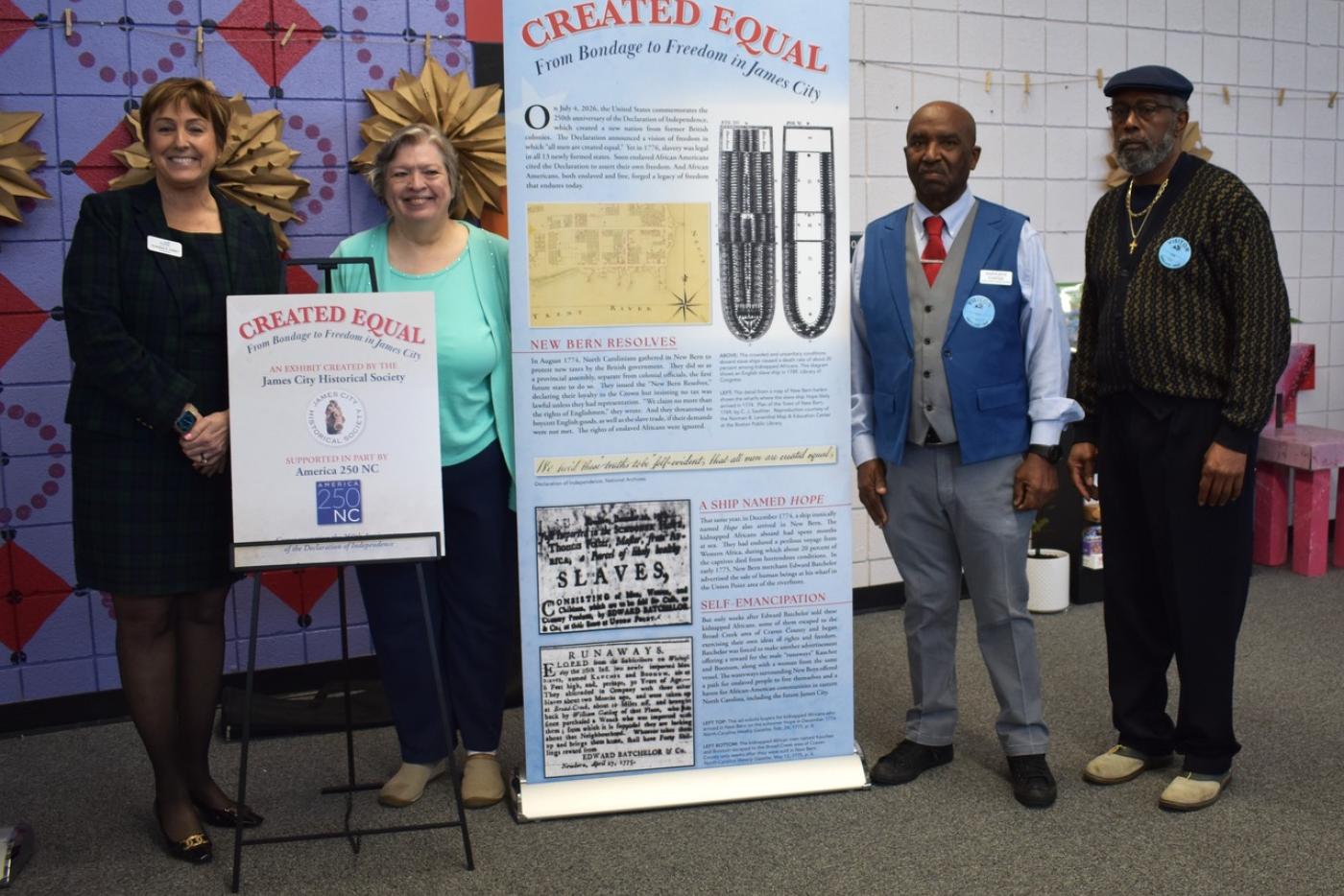 Four people stand beside a display for the exhibit Created Equal: From Bondage to Freedom in James City, created by the James City Historical Society and supported in part by America 250 NC. The group poses indoors with the exhibit banner and a sign announcing the partnership.