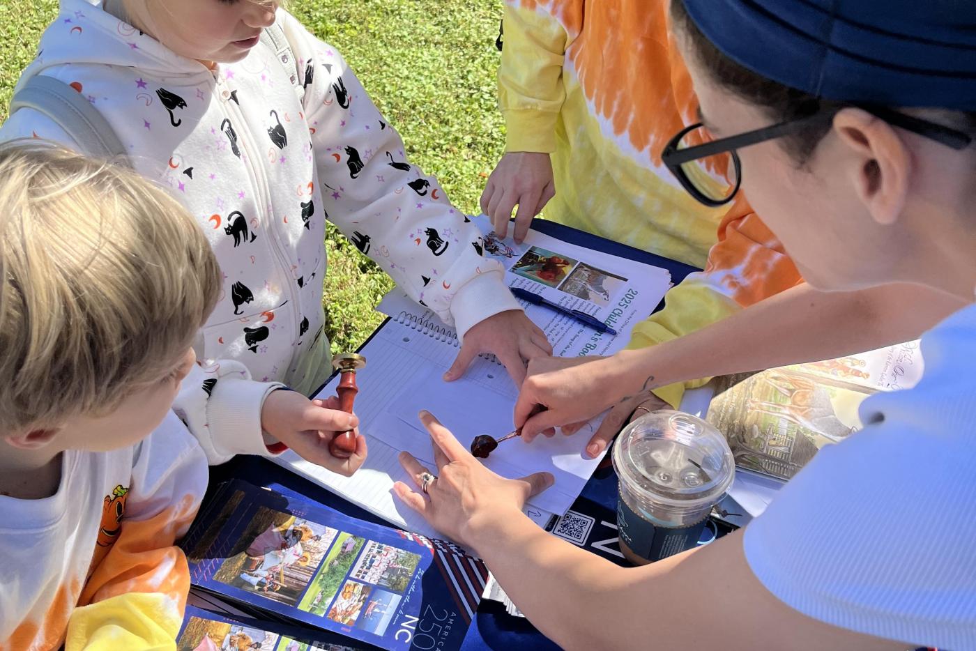 Two women watch as two young children look over a worksheet together. One of the women has her hands on the paper, applying wax. The table has a blue table cloth and various educational materials.