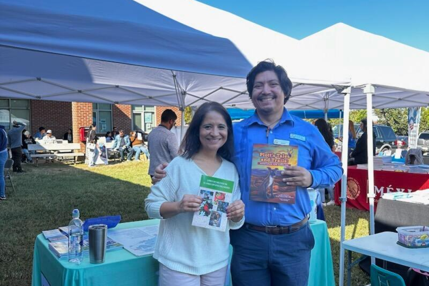 Two people stand in front of a white tent and a table with a teal table cloth. They are holding up books.