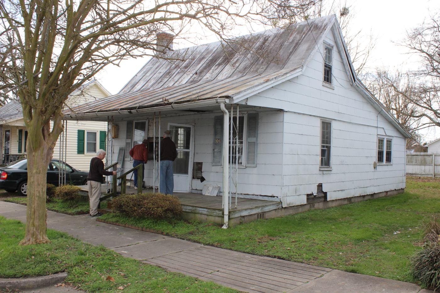 A small, weathered white house with a rusted metal roof and sagging front porch sits on a quiet residential street. Three people stand near the porch examining the structure, while leafless trees frame the scene and neighboring homes are visible nearby.