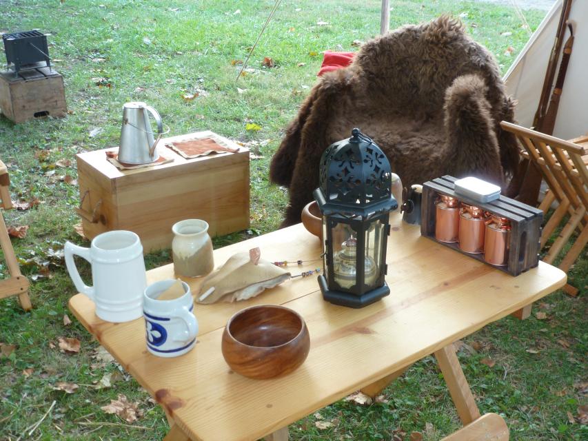 Close up of a camp site table including fur, tent in background, cups, and lantern. 