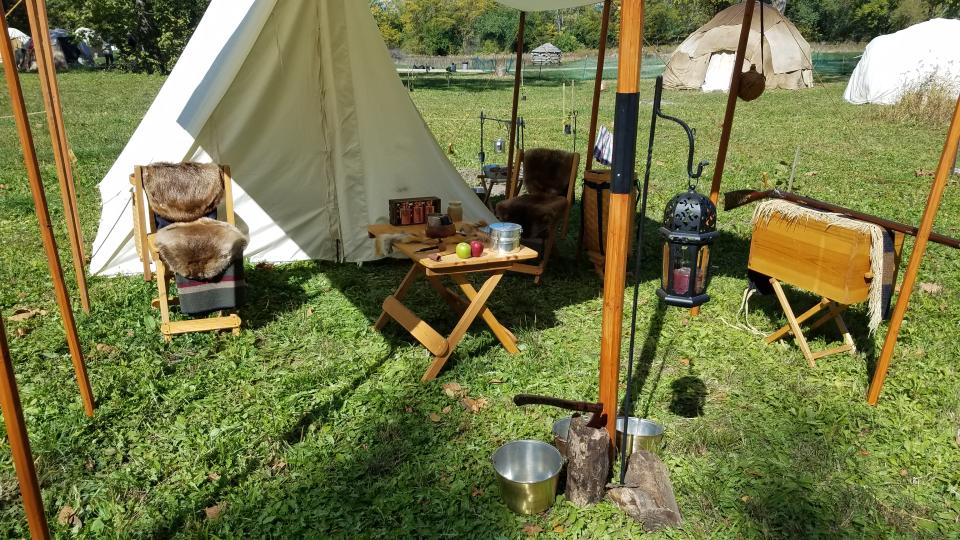 Historic campsite including a white tent, table, and fur covered chair. On grass.