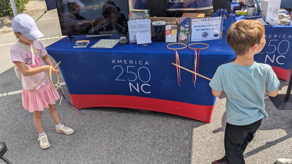 Two young child play Graces by a blue and red America 250 NC tent at an outdoor event.