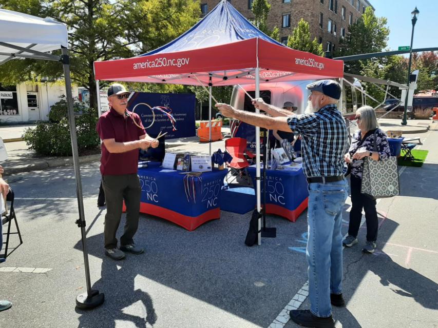 Two men play Graces by a blue and red America 250 NC tent at an outdoor event.