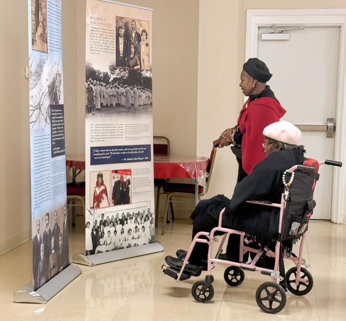 Two women view exhibit panels from Created Equal: From Bondage to Freedom in James City. One woman stands beside another who is seated in a pink wheelchair. Both are dressed warmly, and the panels display historical photos and text about the James City community.