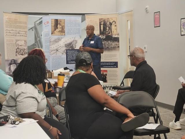 A man speaks to a seated group during a presentation about the Created Equal: From Bondage to Freedom in James City exhibit. Behind him are display panels featuring historical photographs and stories about the James City community’s history.