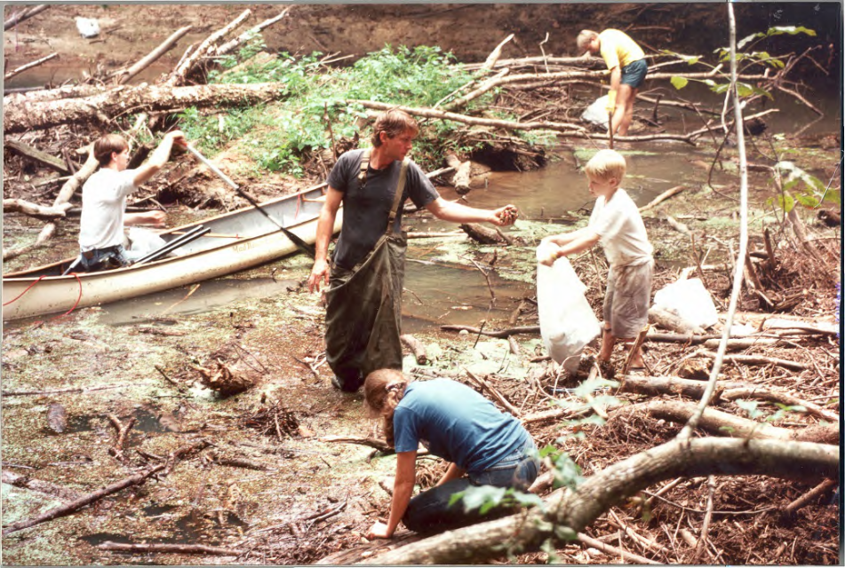A group of people clean a littered creek. One person stands in knee-deep water, handing a piece of trash to another. Others gather debris near a canoe.