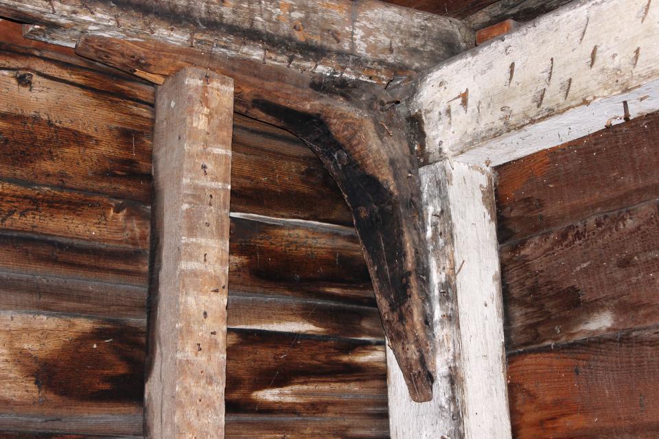 Close-up of the interior wooden framing of an old house, showing dark aged timber walls, exposed beams, and a curved wooden brace supporting the corner structure. Numerous old nails and signs of wear are visible throughout the wood.
