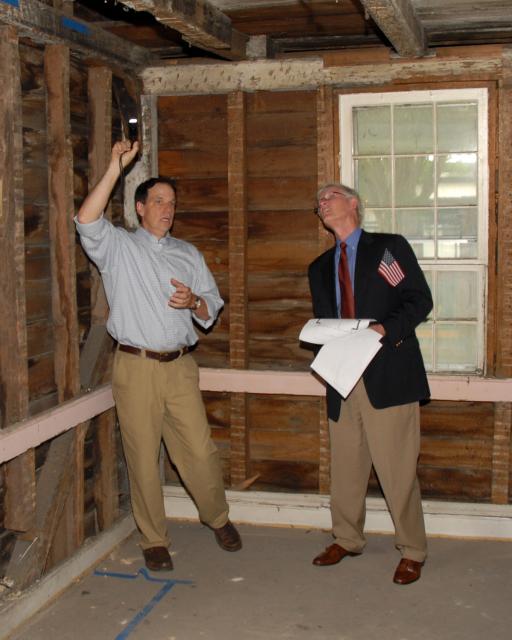 Two men stand inside a stripped-down historic house with exposed wooden walls and ceiling beams. One man shines a flashlight upward while the other holds papers and looks toward the ceiling, appearing to inspect or discuss the building’s structure.