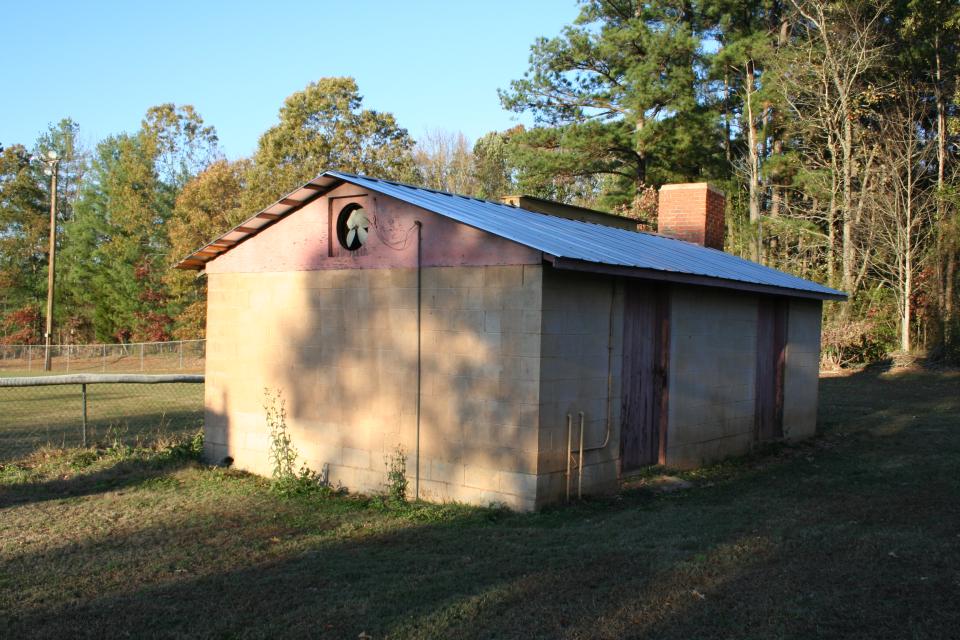 Small cinder block building with a metal roof and wooden doors, surrounded by grass and trees.