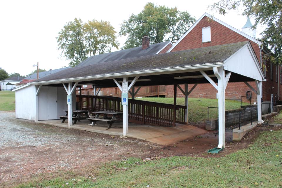Covered outdoor pavilion with picnic tables, white support posts, and an accessible ramp beside a red brick building.