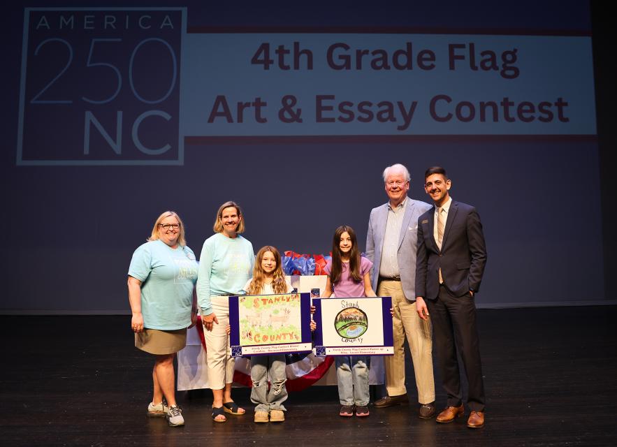 A group of adults and students stand in front of  a screen with the America 250 NC logo