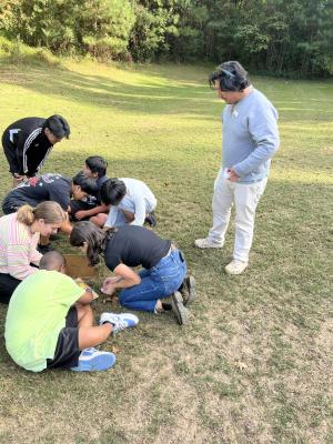 Children gather around papers on the grass outside while an adult looks over their activity