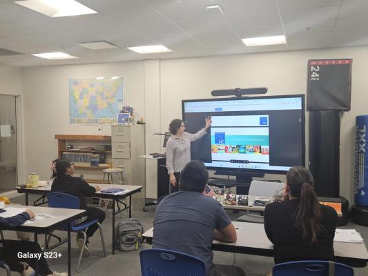 A woman speaks in front of a group of people in a classroom setting. She is facing a large screen and pointing to content. 
