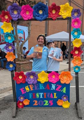 Two people stand behind a selfie frame at the Esperanza Festival. Each hold a book featuring the North Carolina Plott Hound.