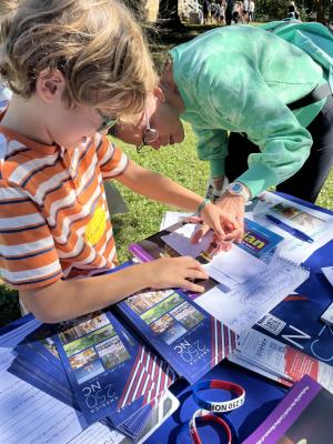 Adult and child work on a wax seal, at a blue table that has an assortment of America 250 NC materials scattered around