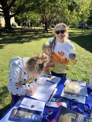 An adult stands behind the America 250 NC table while a child writes on a worksheet. The blue table has America 250 materials scattered around.