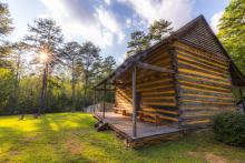 The Allen House, a log dwelling at Alamance Battleground, sits on green grass with tall pine trees surrounding it.