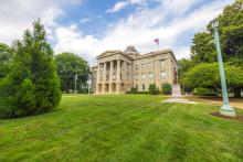 View of a historical building sits on a green lawn with manicured trees in front of it. In front of the building is a statue.