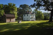 A small cabin, a well and the historic Harper House sit on a green lawn surrounded by old oak trees.