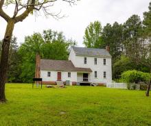 A white house with a brick chimney set in a lush yard with scattered trees under a clear blue sky.