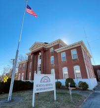 Large brick building in front of blue sky with green grass and a USA flag flying in the front