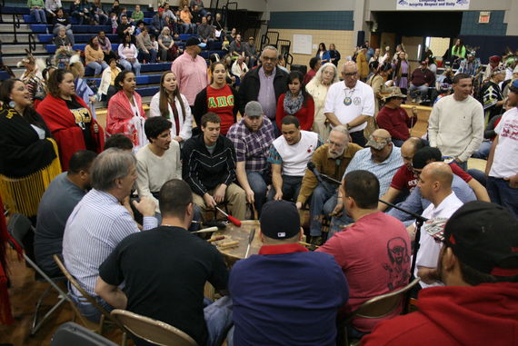 Native group standing around powwow drum in gymnasium.