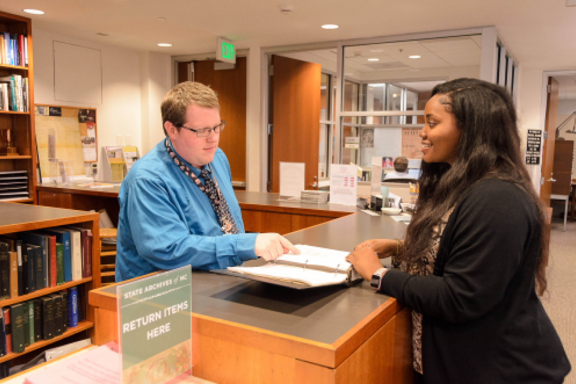 Intern standing to the right of a table across from an archivist in the search room.