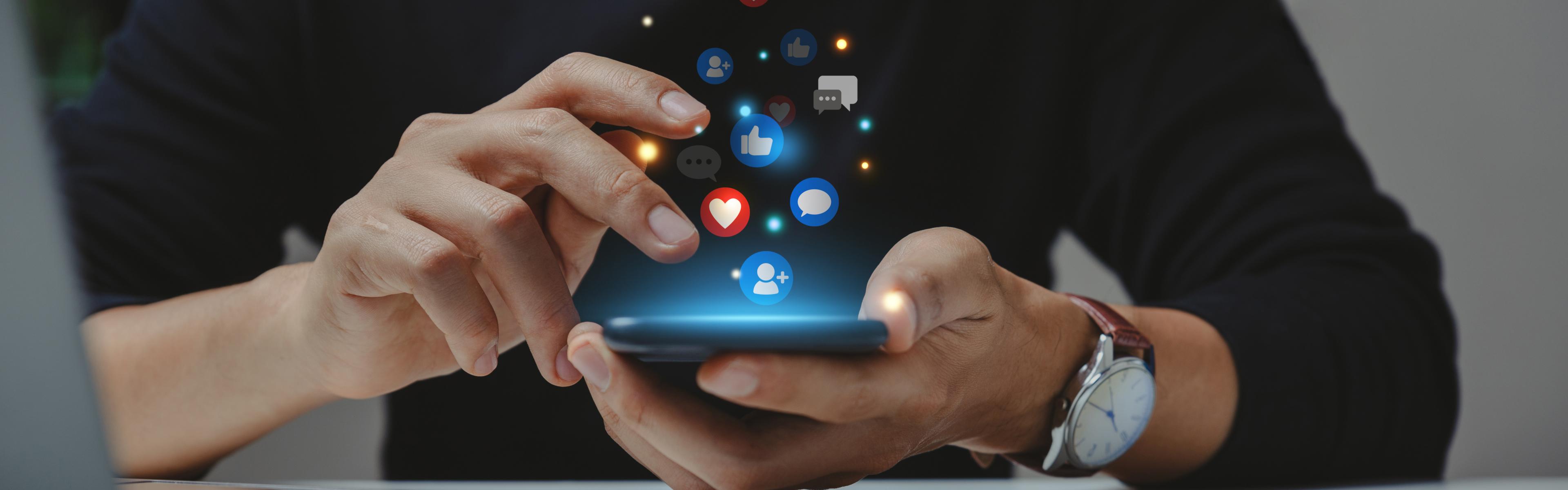 A person sits behind a desk. In their hands they hold a cell phone with images of various social media floating about it. 