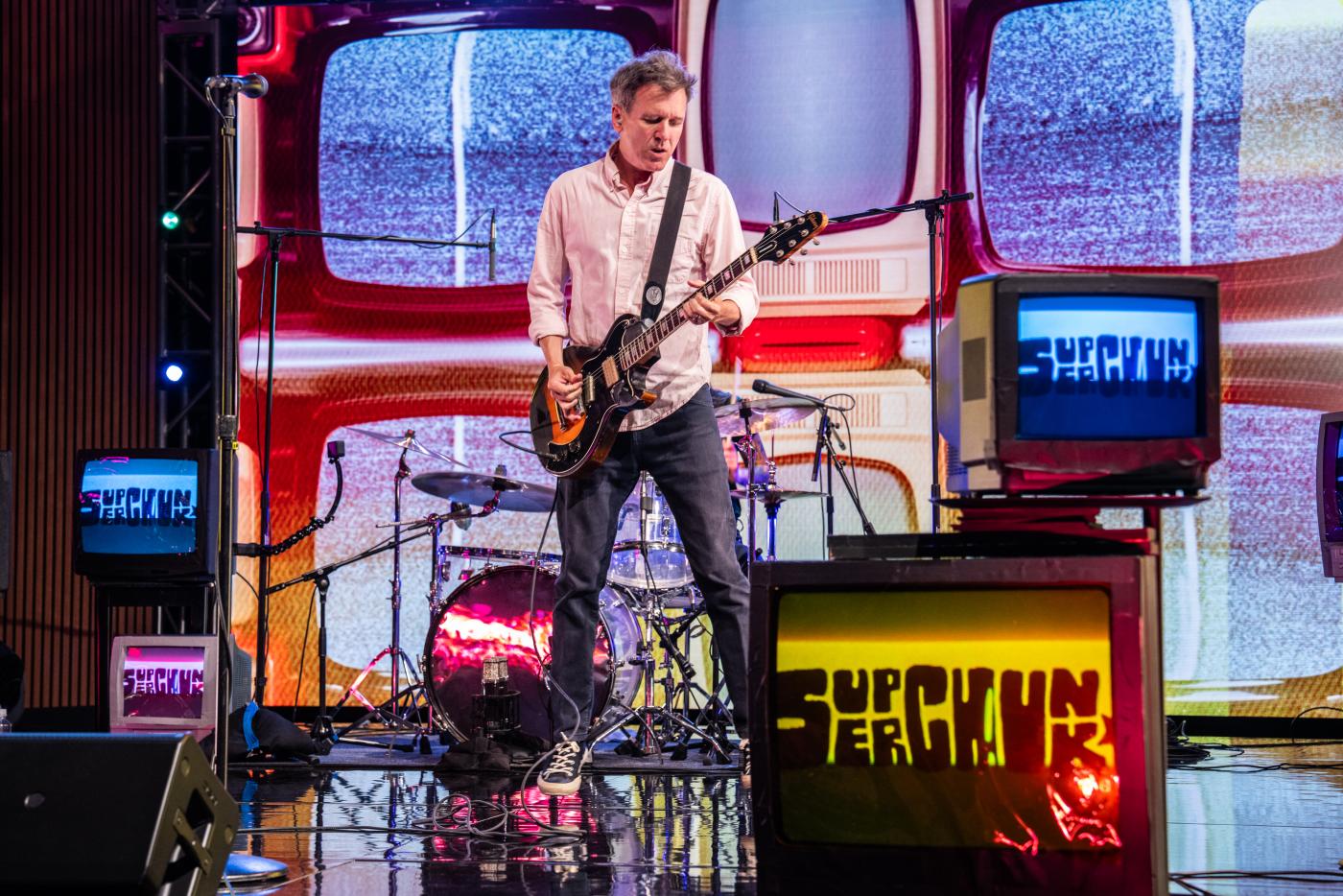 Mac from Superchunk plays guitar in front of a backdrop of static-filled tv screens
