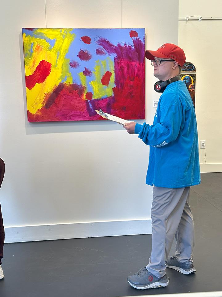 Guy with a baseball hat and headphones around his neck looks at a piece of abstract art. Photo by Eileen Bagnall courtesy of Arts Access
