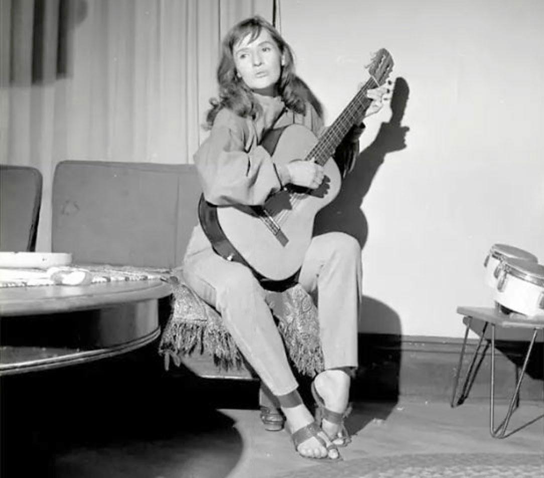 Rosalie Sorrels in black and white, playing guitar in Utah living room in the 1960s.